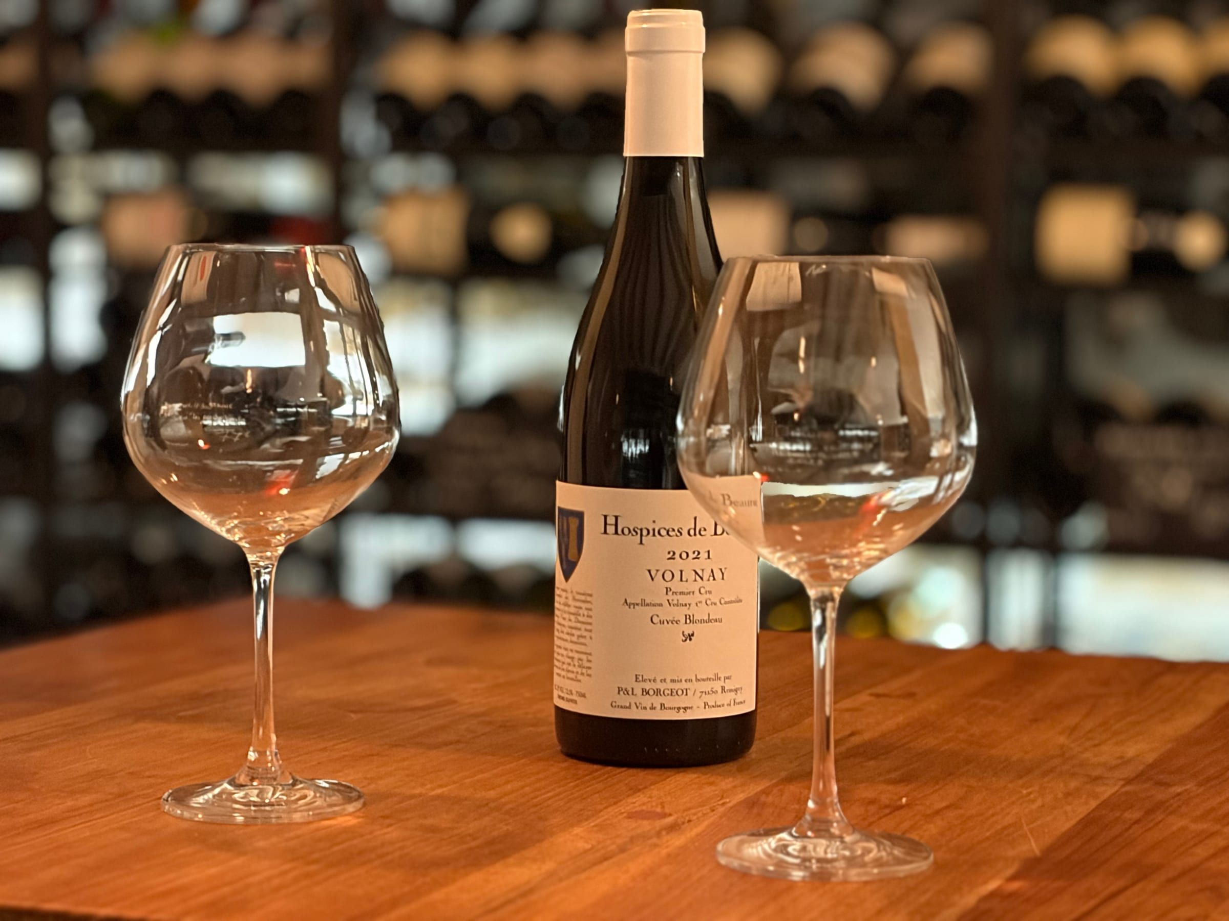 Two wine glasses and a bottle of Volnay Premier Cru Hospices de Beaune on a wooden table