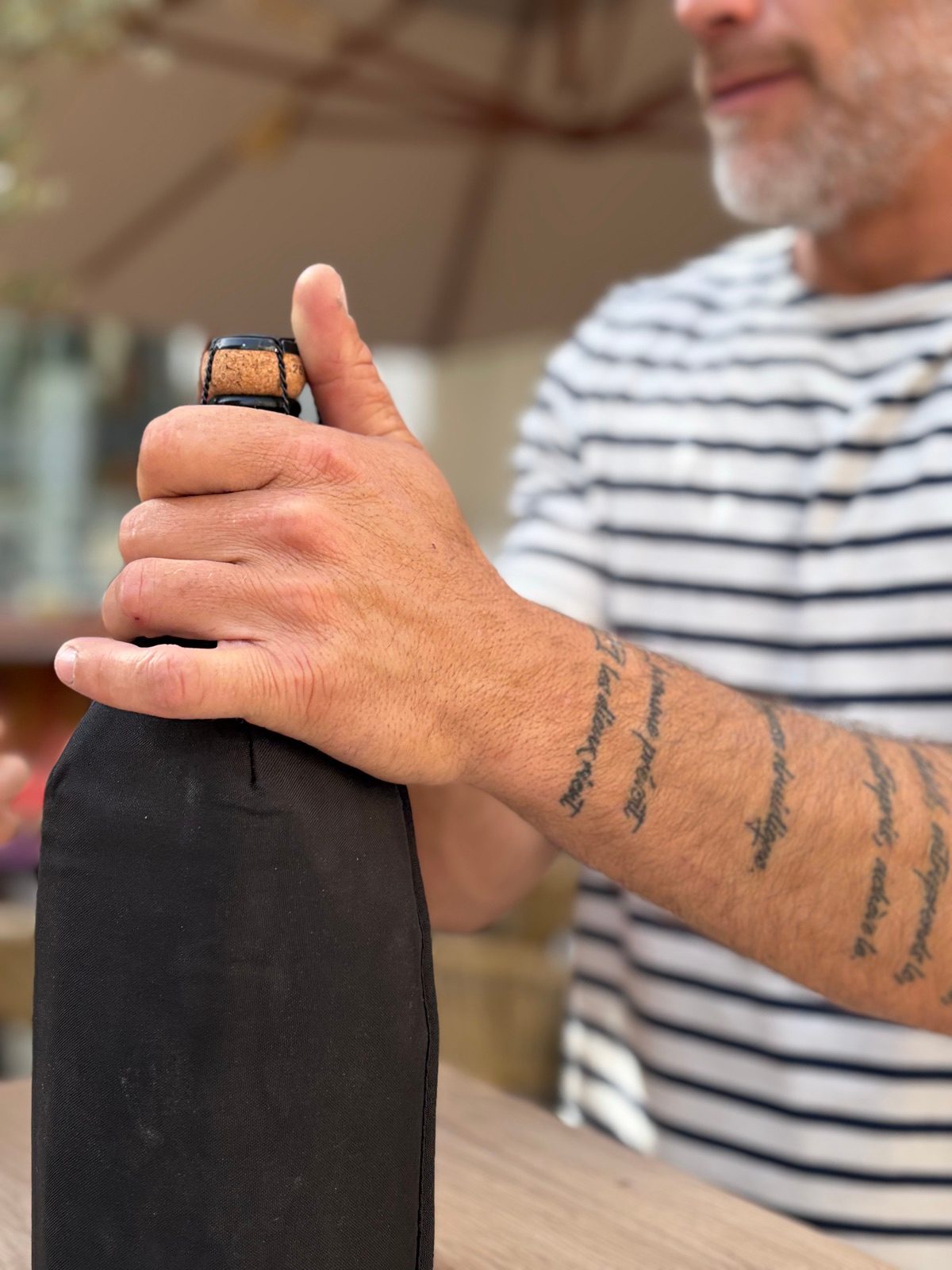 Ben, the wine merchant of L'Oenothèque, in a Breton striped top, tattooed forearm, hand on a bottle he is about to pour